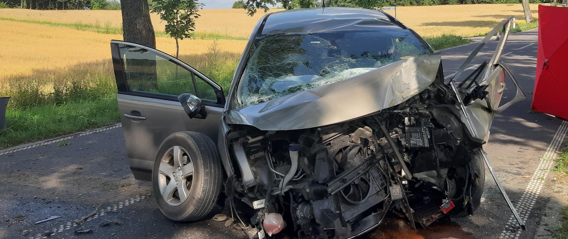 Na drodze stoi z rozbitym przodem samochód osobowy, po jego lewej stronie widoczny jest czerwony parawan. 