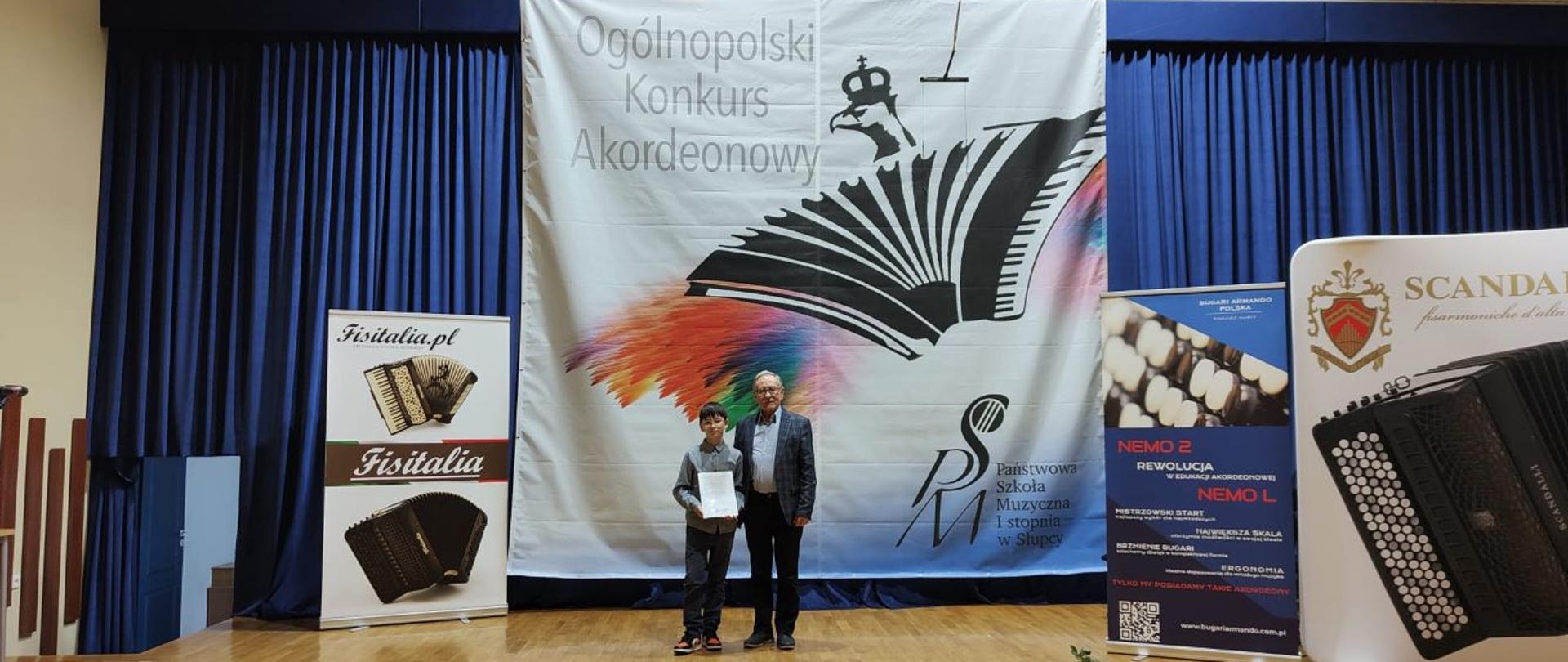 Zdjęcie kolorowe. Chłopiec i mężczyzna stojący na scenie przed dużym banerem z napisem "Ogólnopolski Konkurs Akordeonowy" oraz logo Państwowej Szkoły Muzycznej I stopnia w Słupcy. Chłopiec trzyma w dłoniach dyplom. Mężczyzna w okularach, ubrany w ciemną marynarkę i spodnie. Po lewej i prawej stronie banery reklamowe firm produkujących akordeony: Fisitalia, Bugari Armando oraz Scandalli. W tle ciemnoniebieska kurtyna. Podłoga wykonana z jasnych paneli drewnianych.