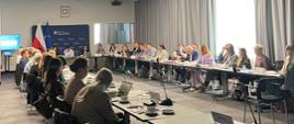 Zdjęcie przedstawia jasną salę. Po lewej stronie w tle widać ściankę Ministerstwa Rozwoju i Technologii. W sali siedzą uczestnicy spotkania przy stołach, które są ustawione w podkowę. Na stołach stoją mikrofony oraz standy z nazwami urzędów.