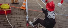 Młody strażak-junior w czerwonym kasku i czarnej koszulce z napisem „MDP”, widziany od tyłu, siedzi na stosie grubych lin na brukowanym placu i aktywnie ciągnie jedną z nich obiema rękami, napinając biały wąż strażacki rozłożony na ziemi. Akcja rozgrywa się podczas zawodów lub pokazu MDP (Młodzieżowych Drużyn Pożarniczych), na co wskazują kolorowe, rysunkowe tarcze celownicze (domek, strażak) w tle, pachołki ostrzegawcze oraz liczni dorośli instruktorzy w piaskowych mundurach strażackich oraz kibice obserwujący wydarzenie. Po lewej stronie, w czerwonym koszu, leży zwinięty żółty wąż. Cały teren jest ogrodzony, a atmosfera jest pełna ruchu i zaangażowania.