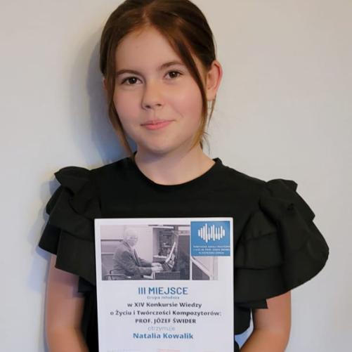 Dziewczynka ubrana na czarno stoi trzymając przed sobą w rękach dyplom.