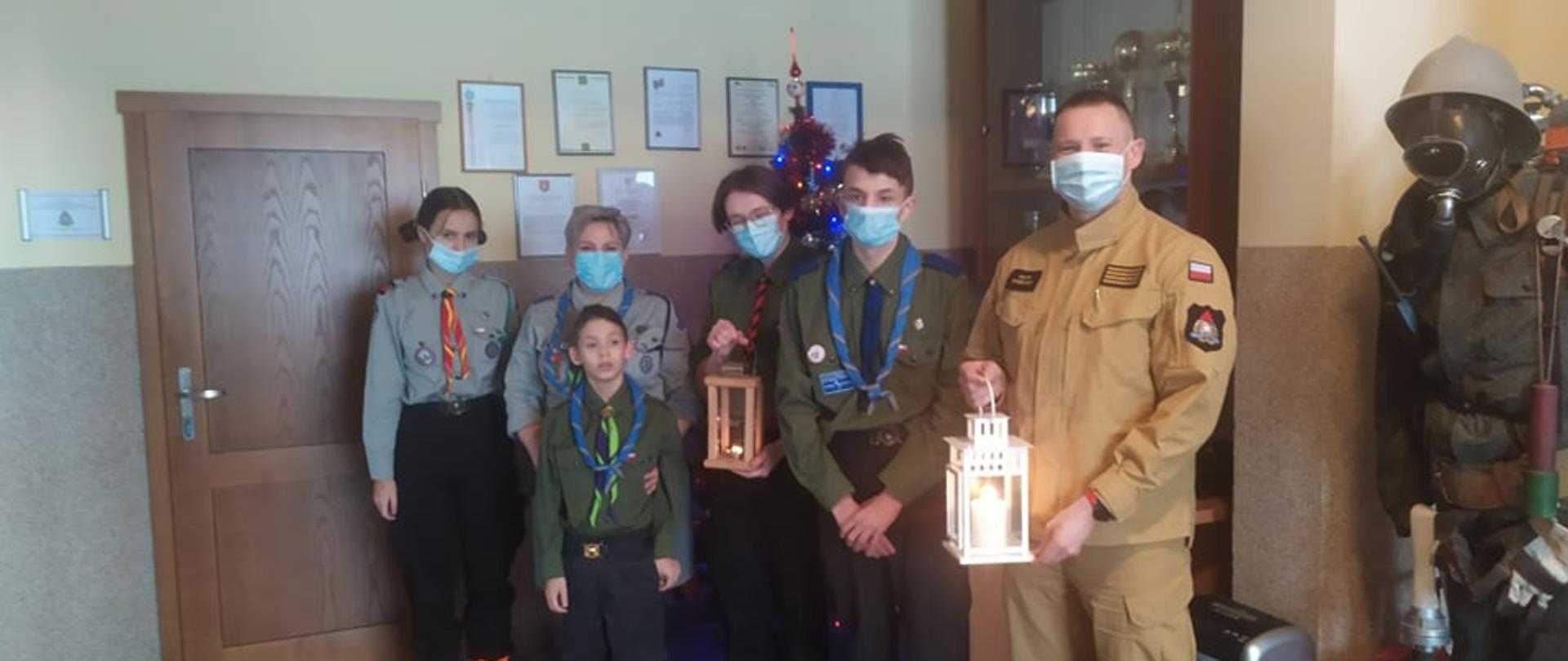 kadr przedstawia harcerzy stojących z funkcjonariuszem PSP. Funkcjonariusz i jeden z harcerzy trzymają w reku lampiony po przekazaniu światełka betlejemskiego.