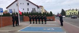 strażacy stoją przed masztem i zostaje podniesiona flaga narodowa na maszt w tle budynki mieszkalne