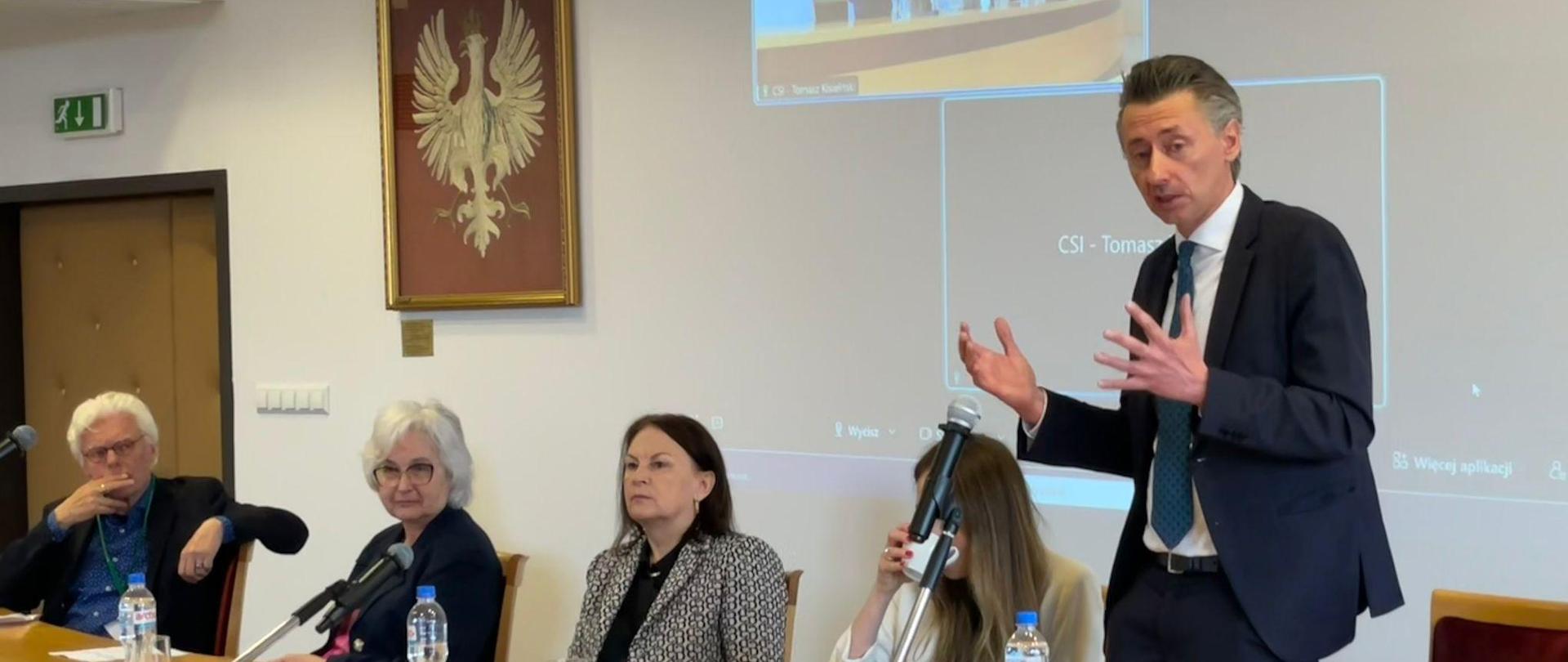 Wiceminister Gdula stoi za stołem, mówi do mikrofonu, obok niego siedzi kilka osób.