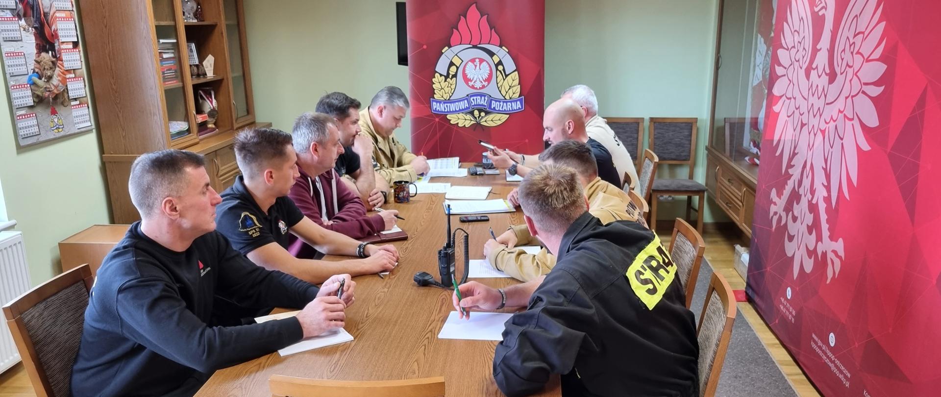 Strażacy siedzą przy stole w sali konferencyjnej. W tle logo Państwowej Straży Pożarnej i wyposażenie pomieszczenia.