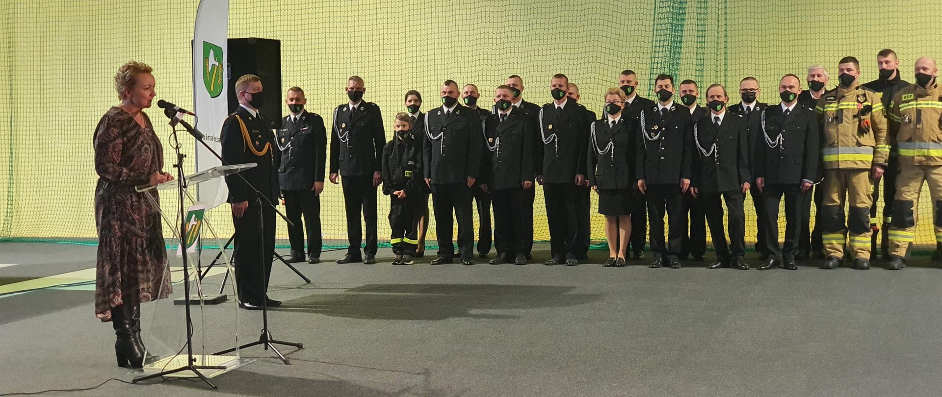 Zdjęcie przedstawia przemawiającą przed mikrofonem Wójt Gminy Barciany oraz druhów OSP ustawionych w dwuszeregu ubranych w czarny mundur wyjściowy lub piaskowe ubranie specjalne.