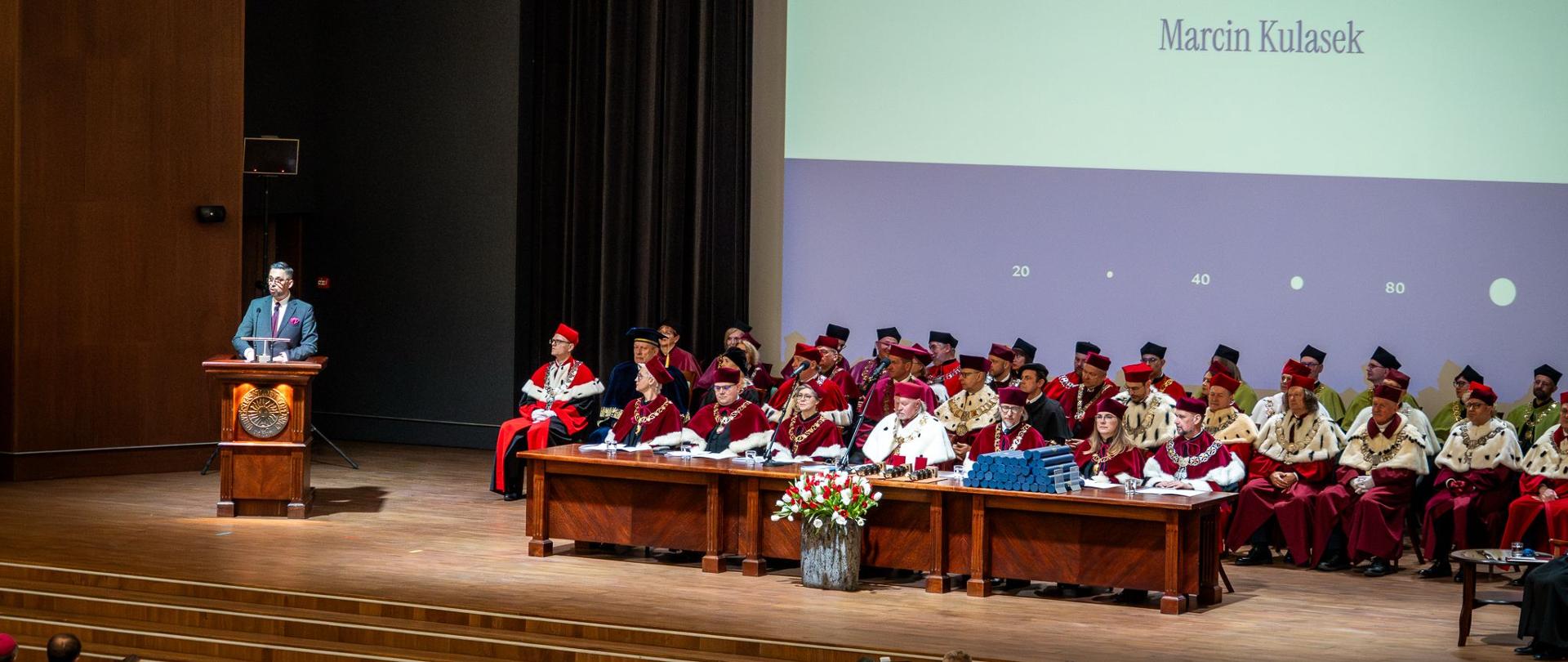 Widok z boku pod kątem na podwyższenie z przodu wielkiej sali, z lewej za mównicą stoi minister Kulasek, pośrdoku i z prawej na krzesłach siedzą ludzie w czerwonych ceremonialnych strojach, za nimi na ścianie wielki ekran w napisem Marcin Kulasek.