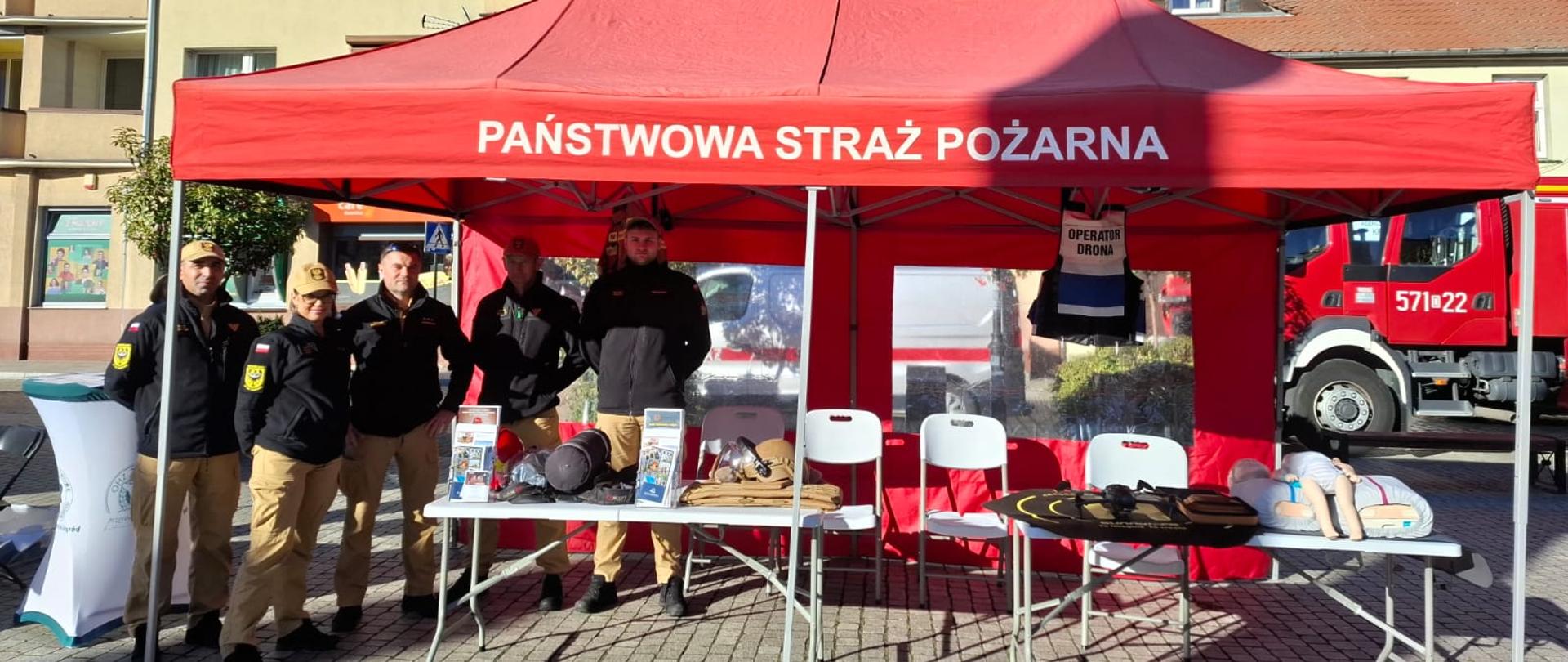 Zdjęcie przedstawia stoisko promocyjne Państwowej Straży Pożarnej usytuowane w słoneczny dzień na miejskim placu wyłożonym kostką brukową.