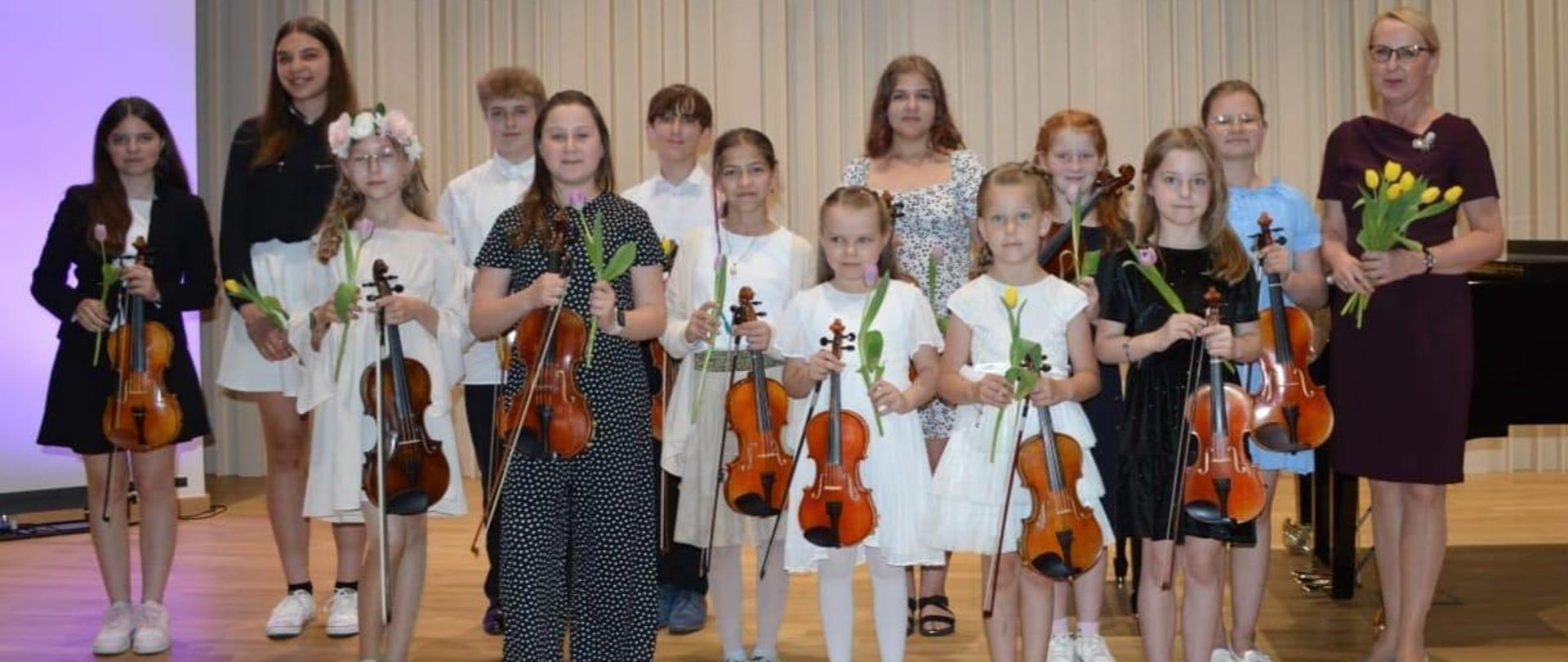 Zdjęcie zostało zrobione podczas koncertu klasy skrzypiec. Na zdjęciu młodzi artyści, którzy zaprezentowali swoje umiejętności gry na skrzypcach. Wszyscy trzymają skrzypce i tulipany, Obok nich kobieta blondynka w okularach w brązowej sukience. 