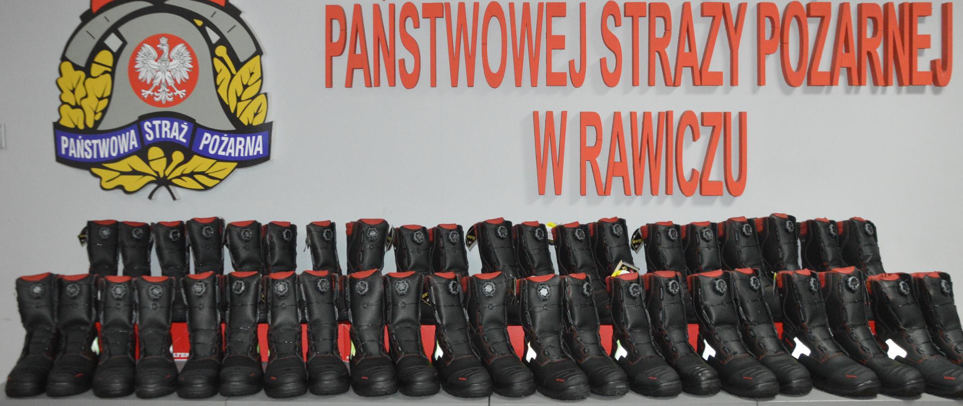 Nowe obuwie specjalne. Świetlica Komendy Powiatowej PSP w Rawiczu. Na stołach, w dwóch rzędach ustawione są nowe buty specjalne - 23 pary. Mają kolor czarny z elementami czerwonymi i żółtymi. W tle na ścianie logo PSP oraz czerwony napis "Komenda Powiatowa Państwowej Straży Pożarnej w Rawiczu"