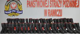 Nowe obuwie specjalne. Świetlica Komendy Powiatowej PSP w Rawiczu. Na stołach, w dwóch rzędach ustawione są nowe buty specjalne - 23 pary. Mają kolor czarny z elementami czerwonymi i żółtymi. W tle na ścianie fragment logo PSP oraz czerwony napis "Państwowej Straży Pożarnej w Rawiczu"
