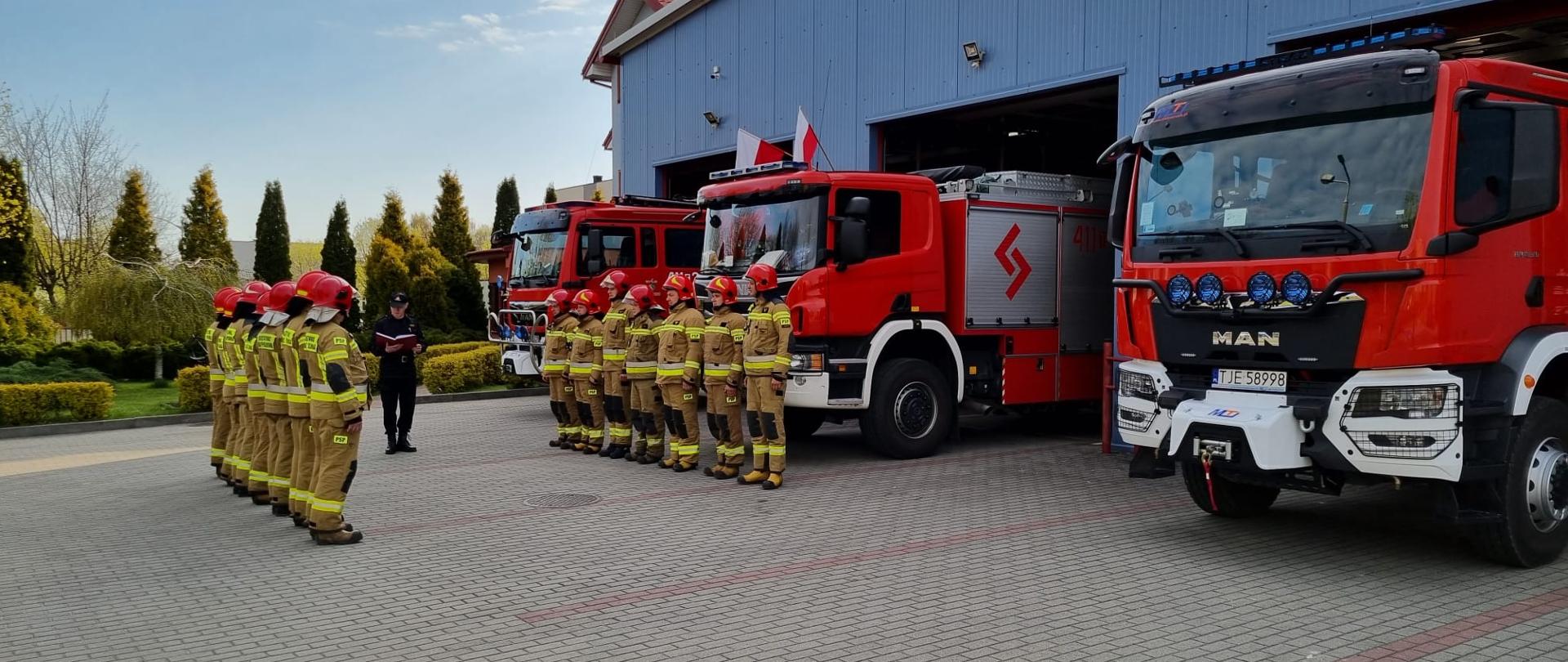 Zdjęcie przedstawia strażaków w umundurowaniu bojowym z założonymi czerwonymi hełmami na głowach. Strażacy podczas uroczystej zmiany służby stoją w dwóch szeregach naprzeciwko siebie. Po prawej stronie znajdują się trzy samochody specjalne. Na drugim planie widnieje strażak w ubraniu dowódczo-sztabowym, który odczytuje rozkaz dzienny.