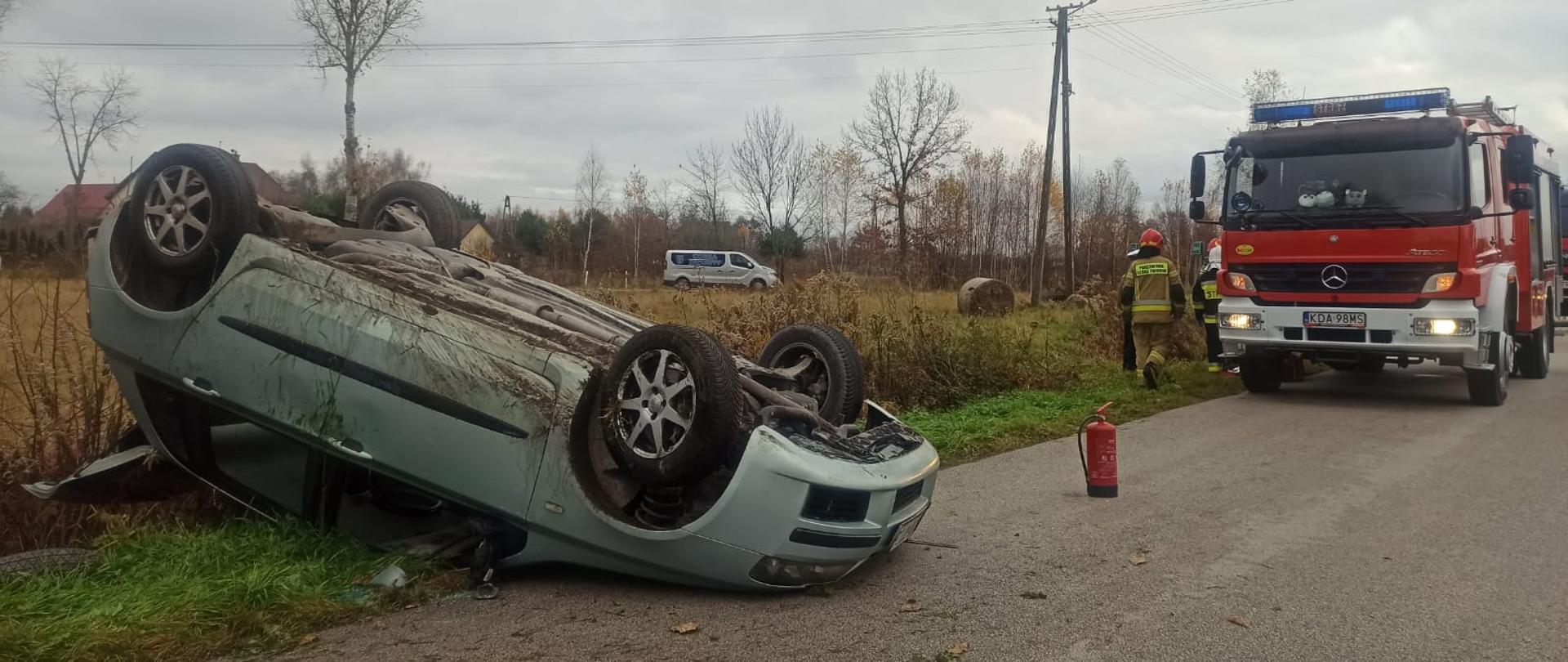 samochód osobowy na jednym pasie ruchu i częściowo w rowie dachował, przed autem gaśnica za pojazdem stoi wóz strażacki i strażacy