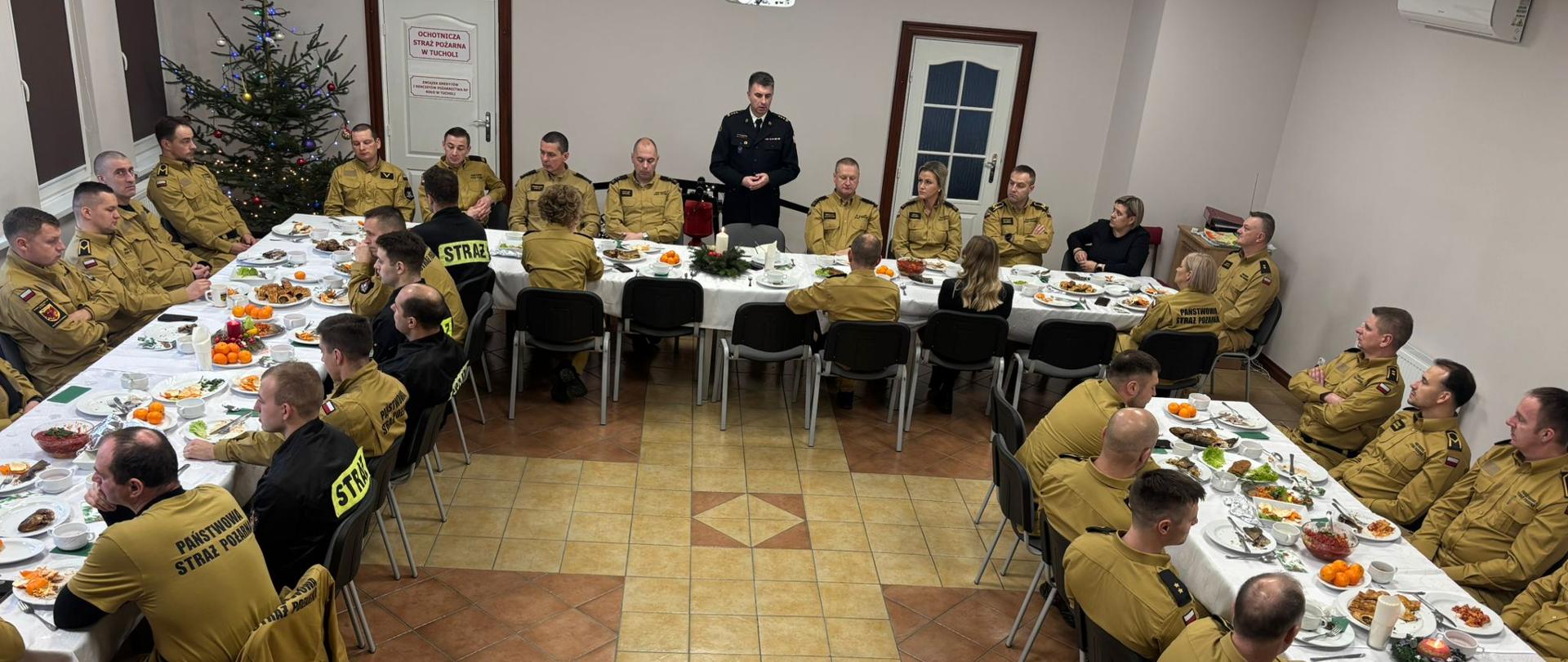 Strażacy znajdują się w pomieszczeniu i siedzą przy stołach. Strażacy są ubrani w mundury w kolorze żółtym. Na środku przy stole stoi Komendant Powiatowy PSP w Tucholi. Komendant ma na sobie mundur galowy w czarnym kolorze. Krzesła są koloru czarnego. Na stołach są białe obrusy. Na stołach znajduje się jedzenie. W lewym rogu stoi choinka. Za choinką jest okno. Obok choinki znajdują się drzwi. Drzwi są kolory białego. Po prawej stronie widać drugie drzwi koloru białego. Ściany są koloru białego. Podłoga jest koloru pomarańczowo-żółtego. 