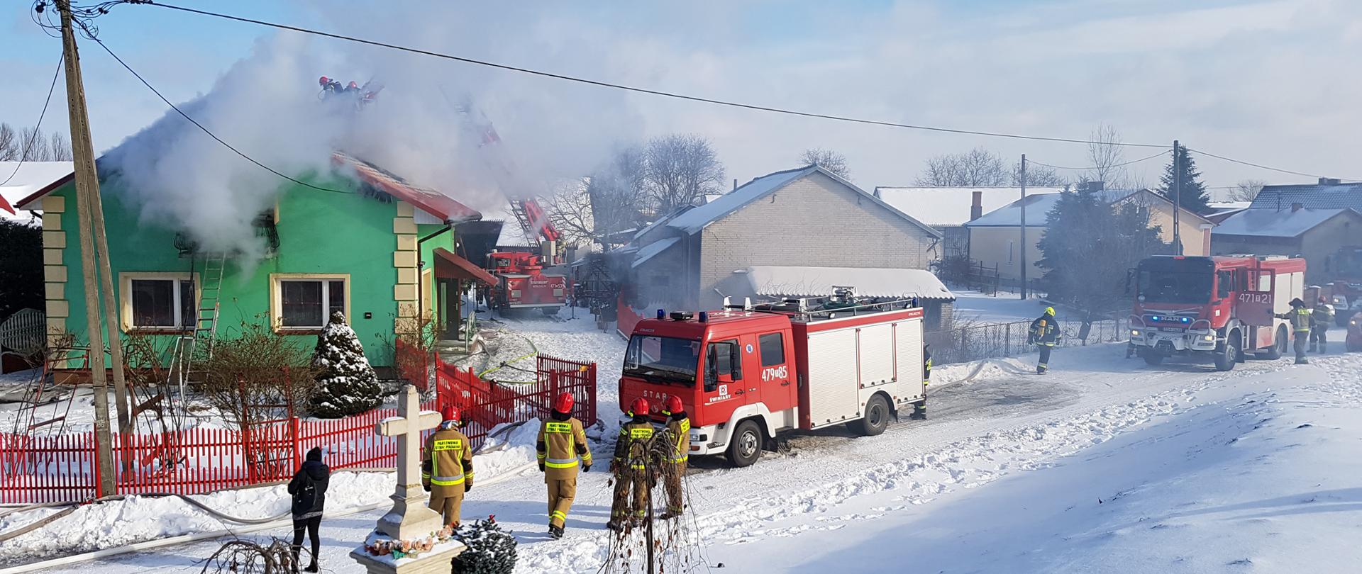 Zdjęcie w zimowej scenerii. Po lewej widać jak z zielonego domu wydobywają się kłęby dymu. Obok domu stoi podnośnik strażacki. W koszu podnośnika jest dwóch strażaków. Po prawej widać budynki mieszkalne i gospodarskie. Na drodze stoją cztery samochody strażackie z włączonymi sygnałami świetlnymi. Jest również siedmiu strażaków, którzy przygotowują się do działań.
