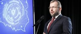 Minister funduszy i polityki regionalnej Grzegorz Puda stoi na scenie przy mikrofonie, po lewej stronie ekran z mapą Polski 