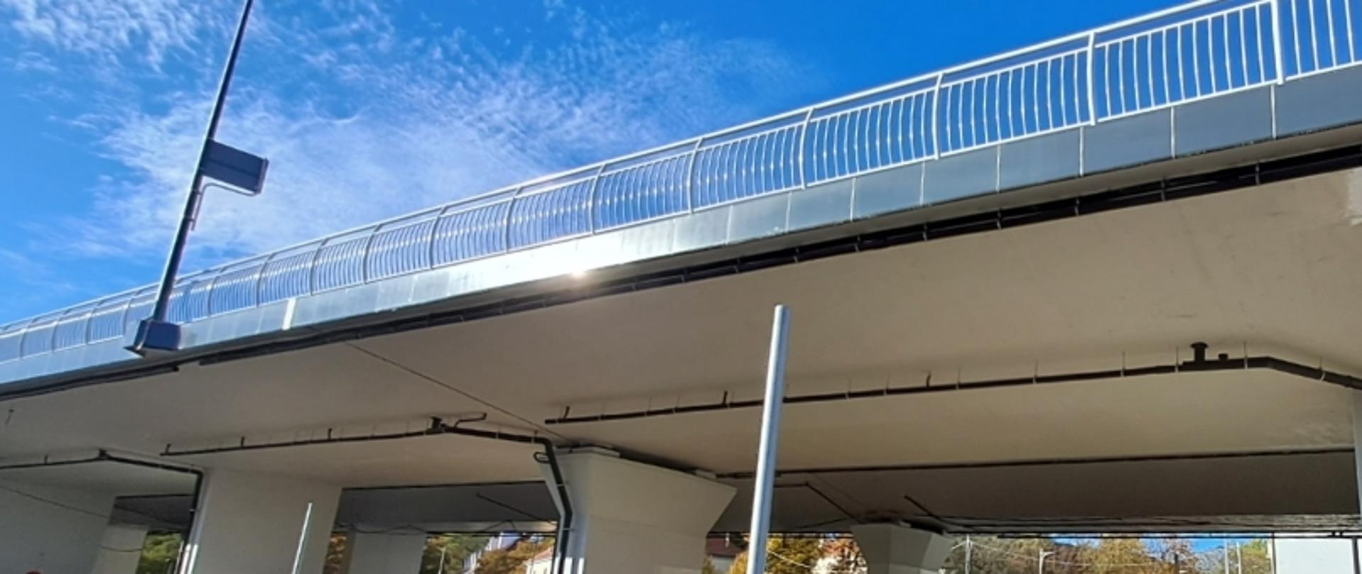 Most lub wiadukt z metalową barierką i latarnią, z widocznymi drzewami poniżej i niebem w tle.