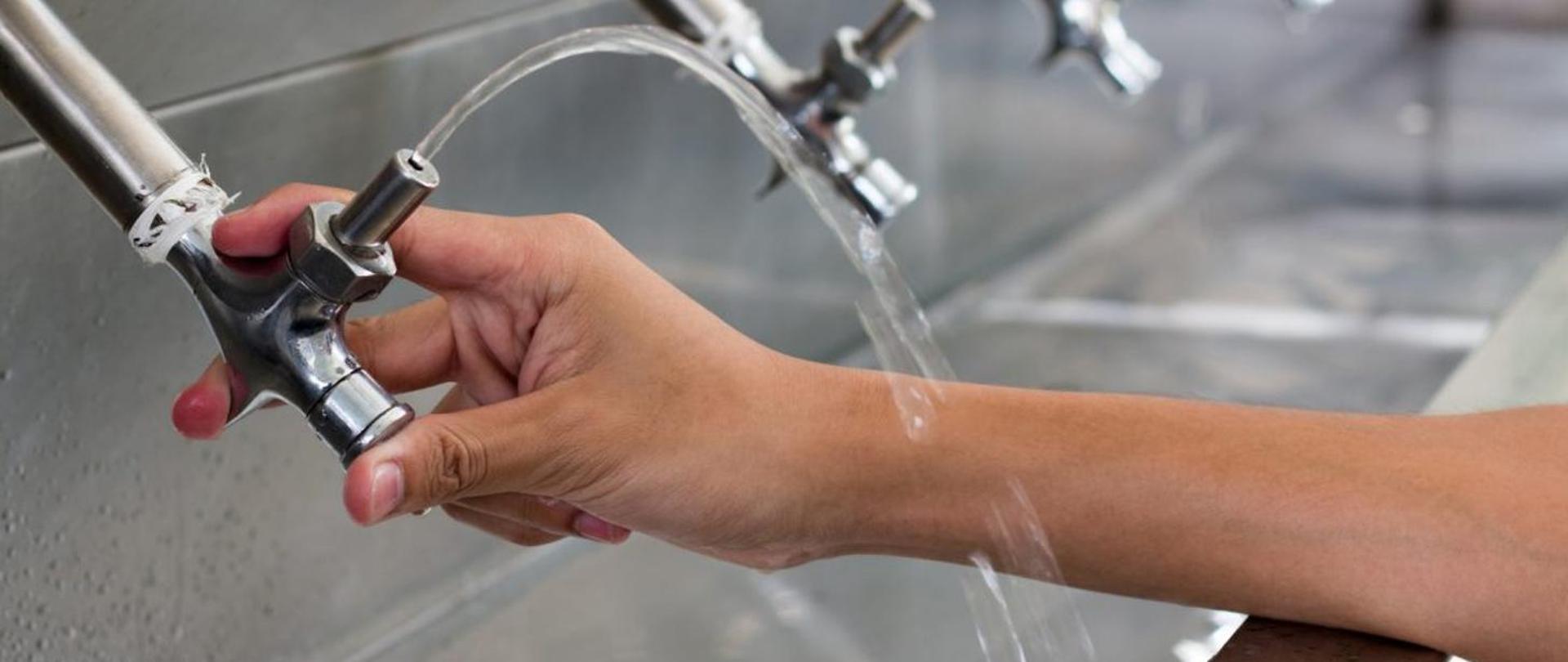 Zbliżenie w perspektywie na trzy łazienkowe dystrybutory wody, przy pierwszym od lewej (najbliższy) ludzka ręka, która go uruchamia.