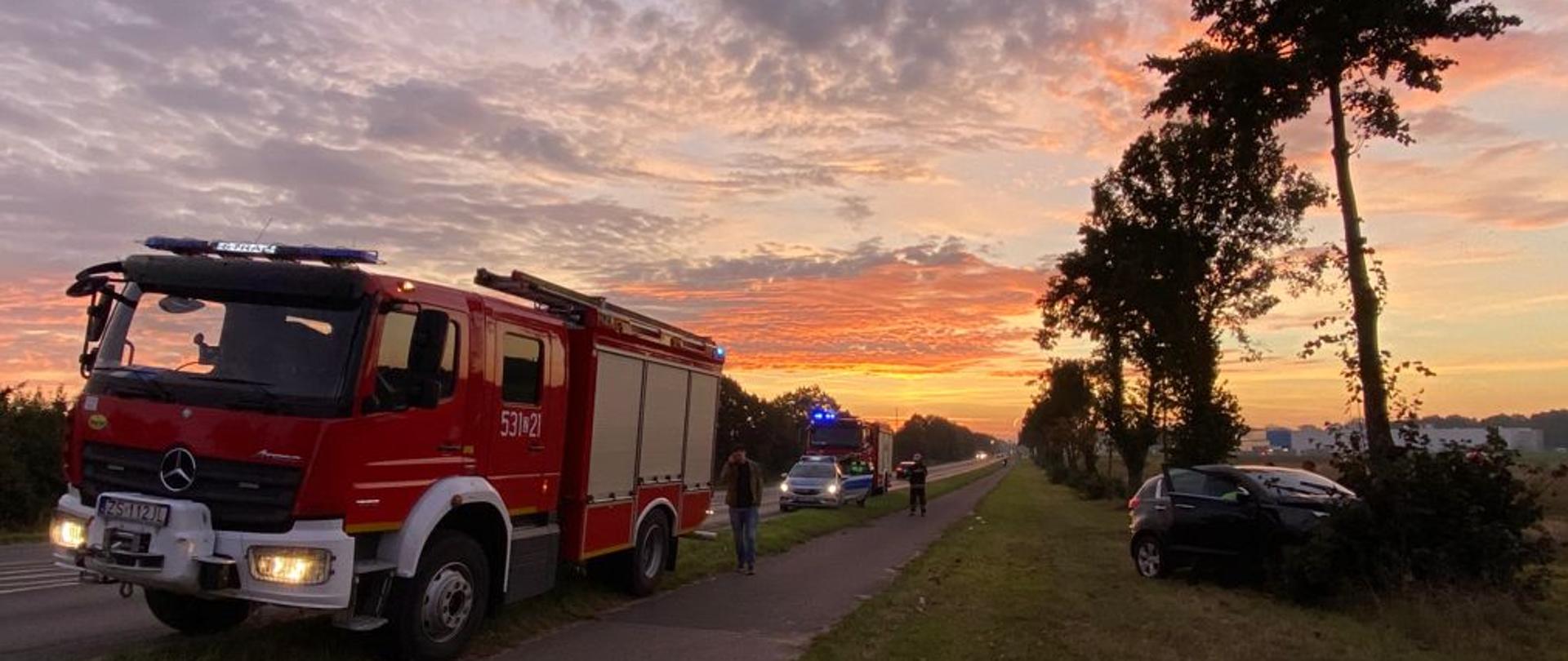 Po prawej czarny samochód uderzył w drzewo, po lewej samochód pożarniczy dalej radiowóz policji i kolejny samochód pożarniczy