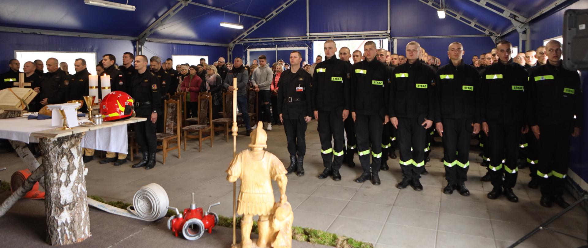 Na zdjęciu widać stojących obok siebie strażaków Zgrupowania Kandydackiego podczas Mszy Świętej obok ołtarz i figura św. Floriana