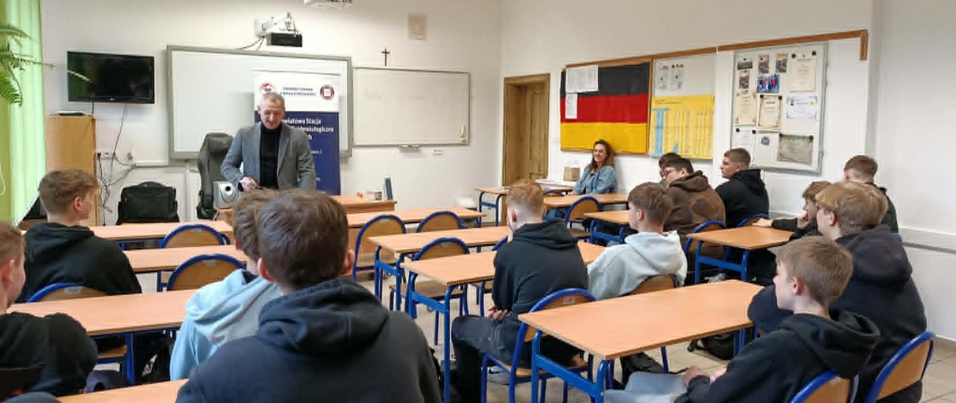 Uczniowie siedzą w klasie, słuchają prelekcji mężczyzny.