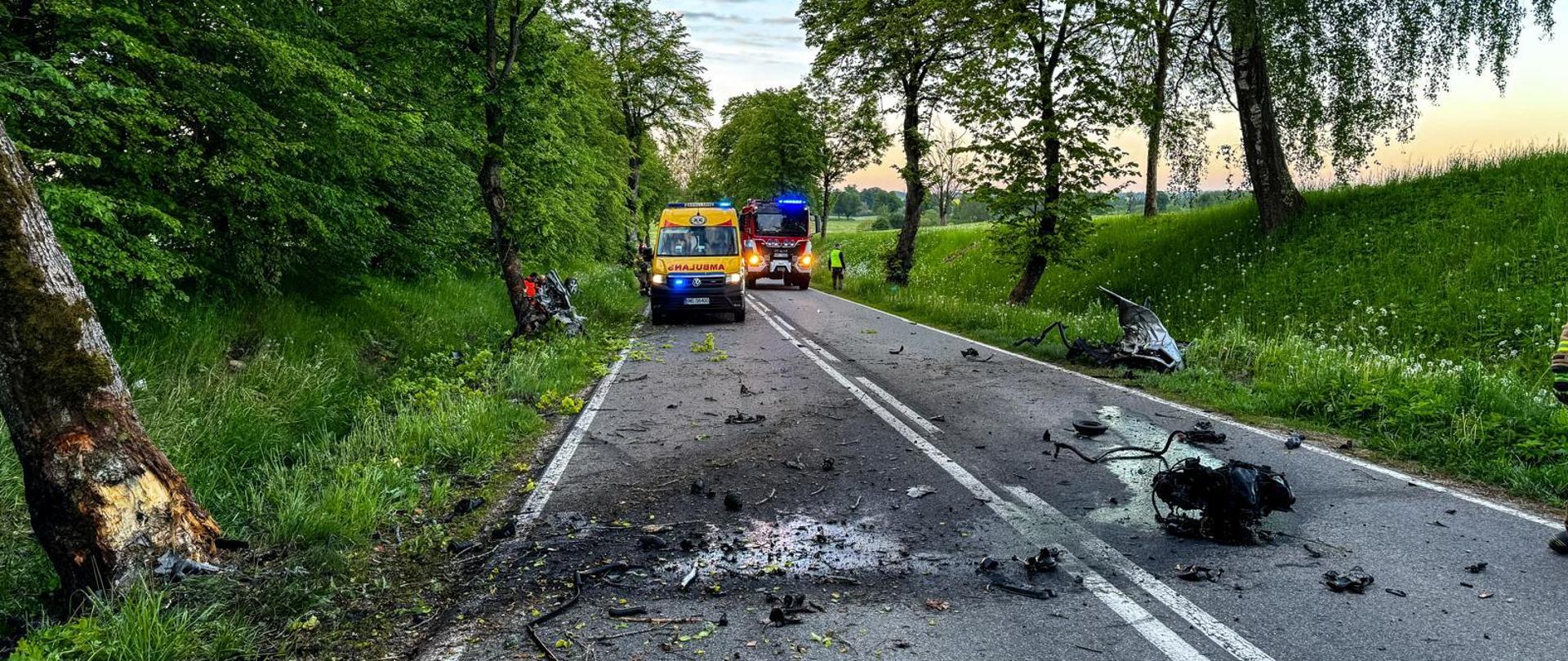 ZRM i straż znajdują się na drodze. Przed pojazdami znajduje się w rowie uszkodzony samochód, na drodze leży wyrwany silnik z pojazdu. 