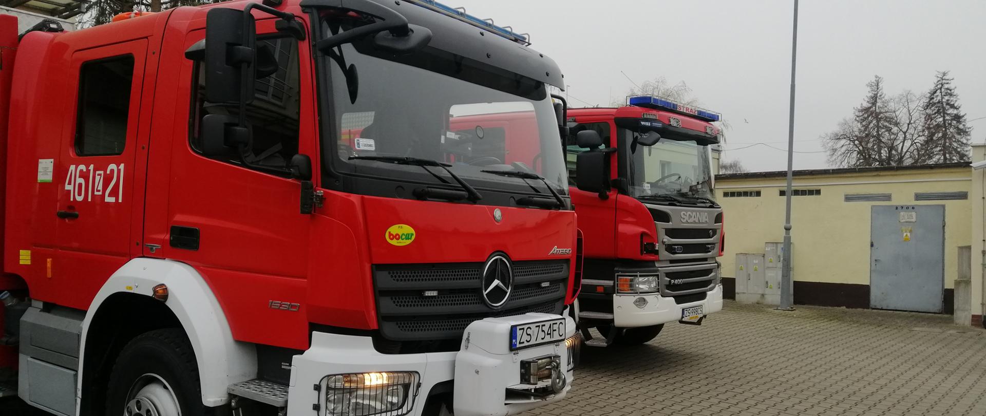 Zdjęcie przedstawia zewnętrzną część kabiny dwóch wozów bojowych które znajdują się na placu jednostki ratowniczej w Kamieniu Pomorskim. W tle widać słup od lampy oraz metalowe drzwi.