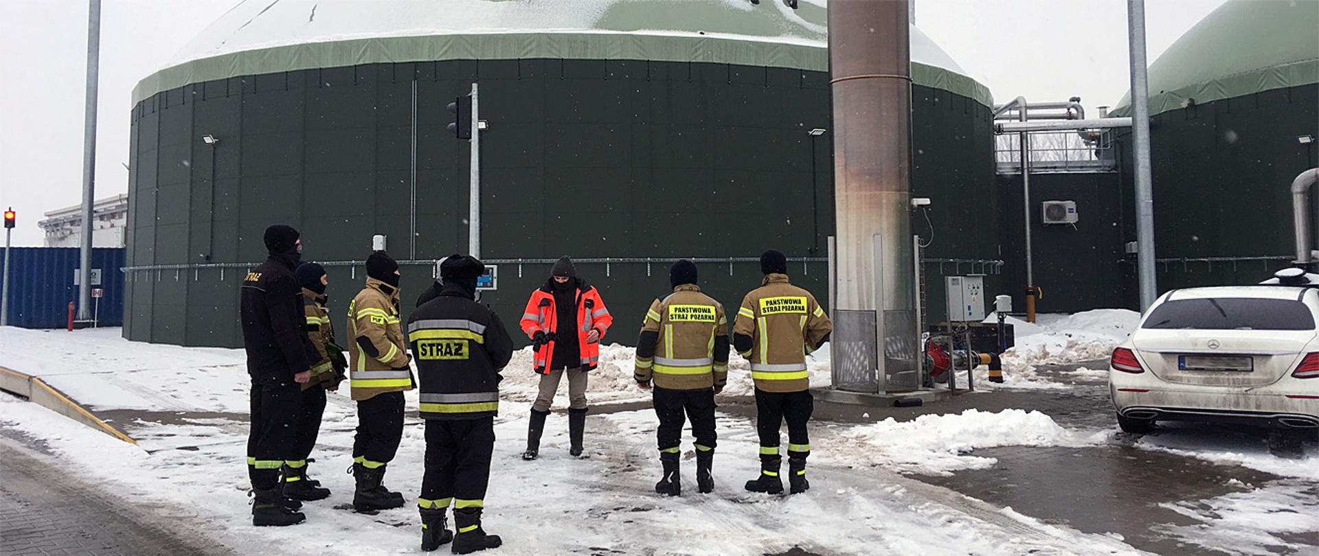 Na zdjęciu widać grupę siedmiu osób, sześciu z nich to funkcjonariusze Państwowej Straży Pożarnej i jedna osoba cywilna. Strażacy ubrani są w ubrania bojowe typu Moratex, na twarzy mają czarne wełniane kominiarki, osoba cywilna ubrana jest w kurtkę barwy pomarańczowej z nakryciem głowy w postaci czapki z pomponem. Strażacy są ubrani w czarne spodnie i kurtki barwy jasnobrązowej z pasami odblaskowymi barwy żółtej i szarej z napisem na plecach Państwowa Straż Pożarna, na nogach mają skórzane buty z wysoka cholewką. W tle widać dwa duże obiekty o kształcie owalnym przypominające silosy barwy ciemnozielonej, połączone ze sobą instalacja rurową. Obok strażaków stoi duży metalowy komin barwy szarej, po prawej stronie fotografii zaparkowane auto barwy białej. Na ziemi leży śnieg, miejscowo odgarnięty.
