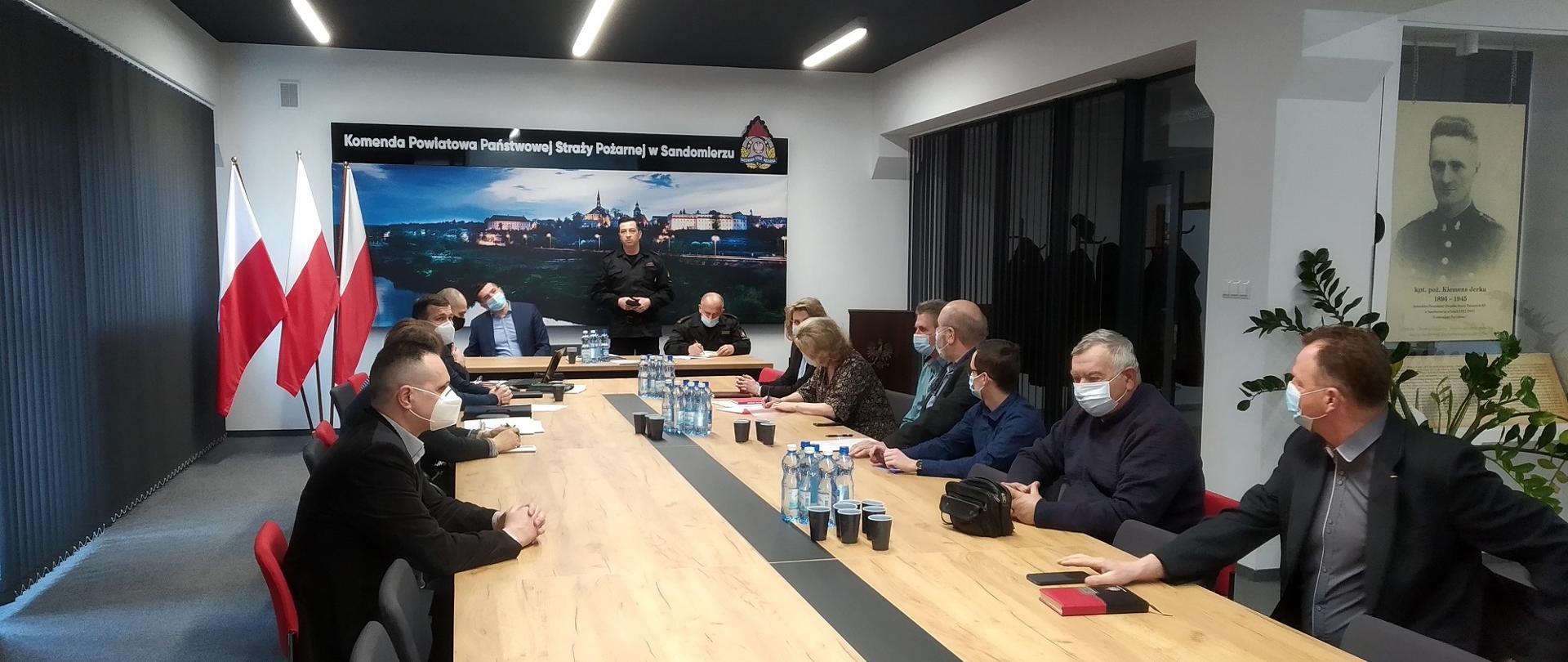 Zdjęcie zrobione w reprezentacyjnej Sali. widać zebranych siedzących przy stole w oddali jedna osoba stoi.