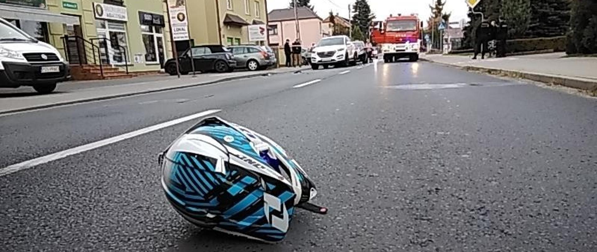 Niebiesko-biały kask motocyklowy leżący na drodze. W tle budynki, samochody osobowe i samochód ratowniczo-gaśniczy.