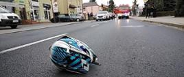 Niebiesko-biały kask motocyklowy leżący na drodze. W tle budynki, samochody osobowe i samochód ratowniczo-gaśniczy.