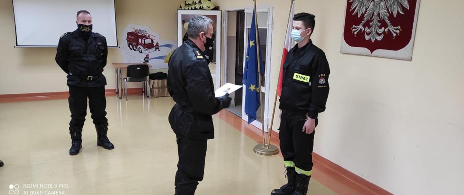 Komendant Powiatowy stojący po środku zdjęcia w mundurze sztabowym koloru czarnego wręcza akt ślubowania nowoprzyjętemu pracownikowi stojącemu po prawej stronie kadru. W tle widoczny fragment godła - orzeł biały na czerwonym tle po lewej stronie ślubującego widoczna flaga Rzeczpospolitej Polskiej oraz Unii Europejskiej. Po lewej stronie przed tablicą multimedialną stoi Zastępca Dowódcy Jednostki Ratowniczo Gaśniczej.