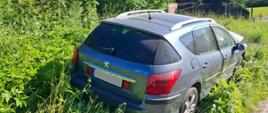 Samochód osobowy Peugeot, koloru szarego stojący na poboczu drogi, które jest porośnięte wysoką trawą.