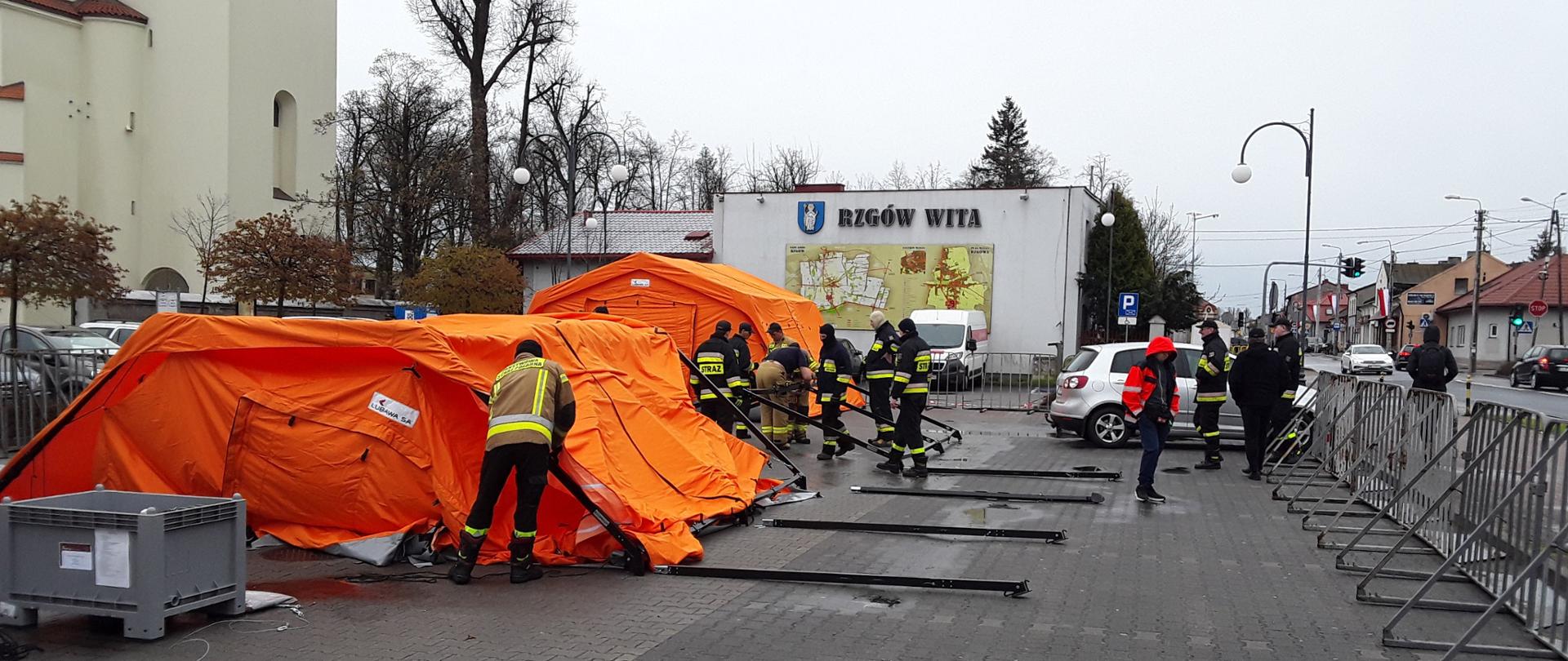 na zdjęciu namioty stelażowe pomarańczowe tworzące mobilny punkt szczepień, strażaków rozkładających te namioty, w tle budynki i ulica