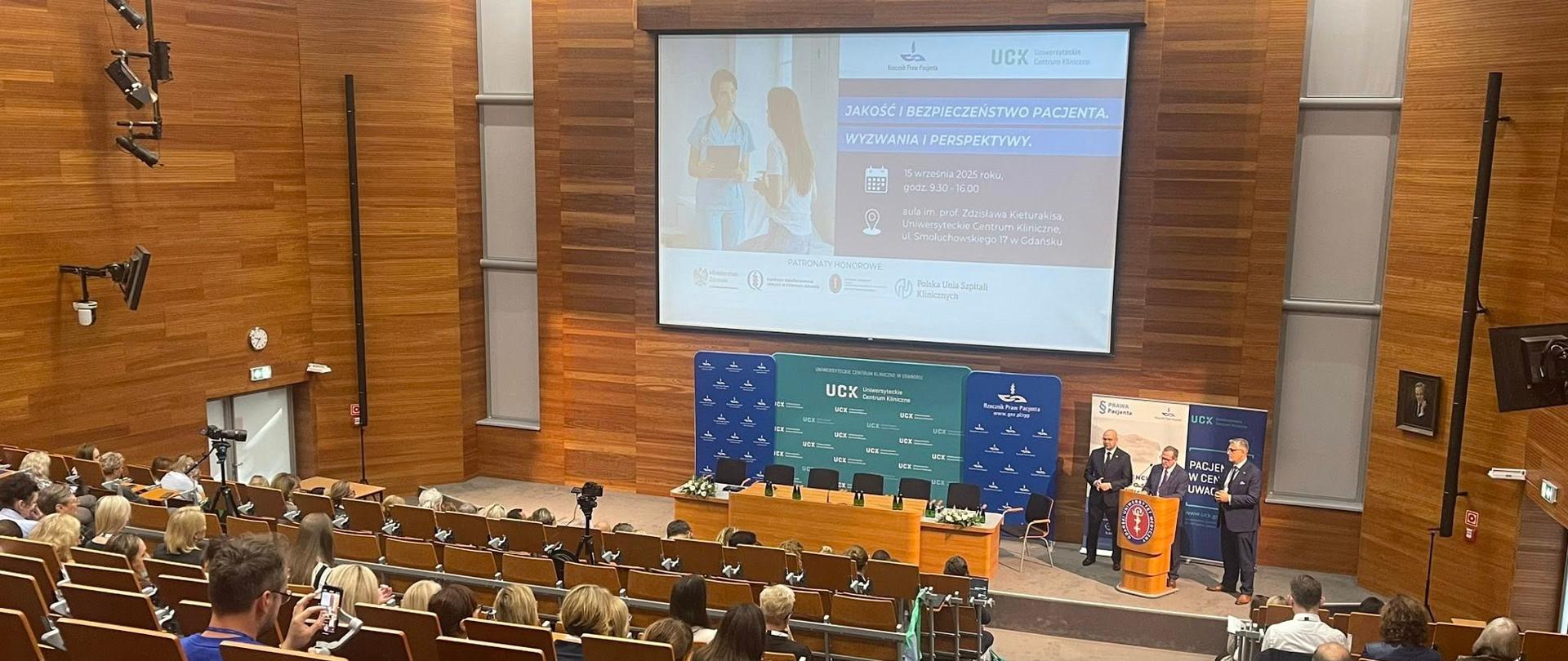 Zdjęcie przedstawia aulę, gdzie widać prezentację oraz osoby przy mównicy otwierające konferencję.