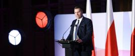 Na oświetlonym podwyższeniu przy mównicy stoi minister Czarnek i mówi do mikrofonu, za nim polskie flagi.