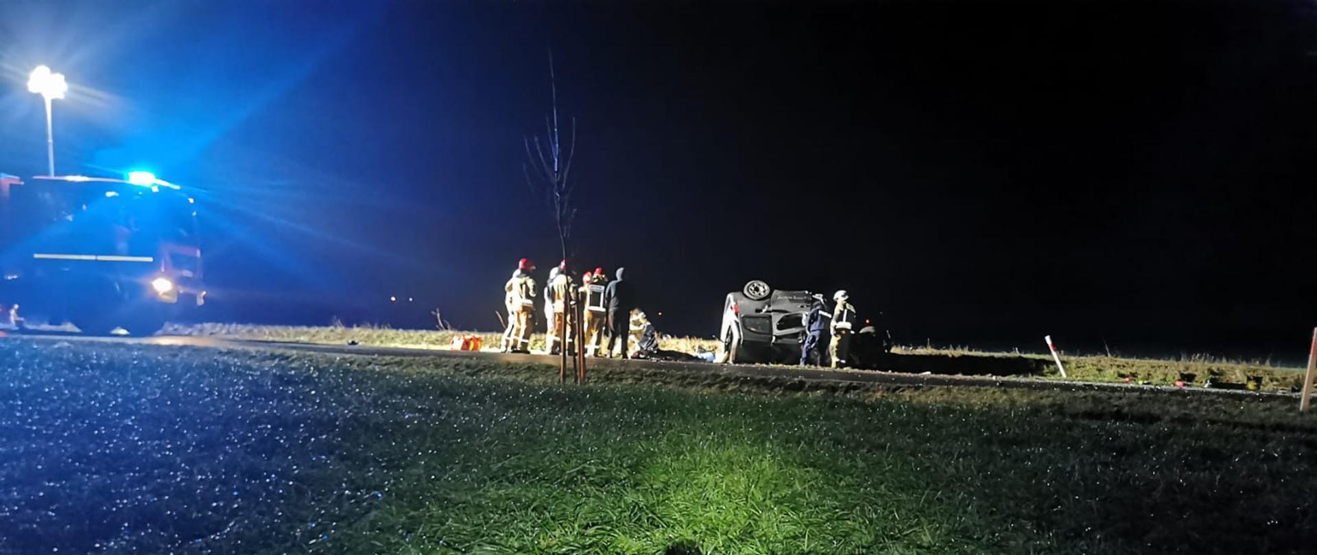 Na zdjęciu wdać silnik który wypadł z auta, a na drugim planie służby ratunkowe przy rozbitym pojeździe 