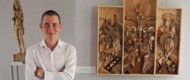 Młoda osoba, stoi, za nią prace rzeźbiarskie w drewnie (trzyczęściowy, niewielki ołtarz oraz, dalej, stojąca postać); patrzy w obiektyw