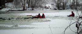 Na środku zamarzniętej rzeki po prawej stronie strażak siedząc na lodzie w ubraniu niezatapialnym, skierowany w lewo, przy pomocy linki wyciąga z przerębla strażaka, który (ubrany w ubranie niezatapialny) wyciągnął poszkodowanego, strażaka ubranego w ubranie niezatapialne z wody tyłem skierowany do ratującego go strażaka, Wszyscy siedzą na lodzie.