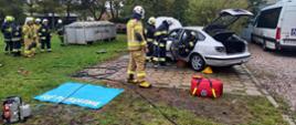 Zdjęcie przedstawia strażaków OSP podczas egzaminu praktycznego podczas którego musieli uratować osobę poszkodowaną z rozbitego samochodu osobowego. Z lewej strony widoczna jest grupa strażaków ratowników OSP obserwujących ćwiczenie.