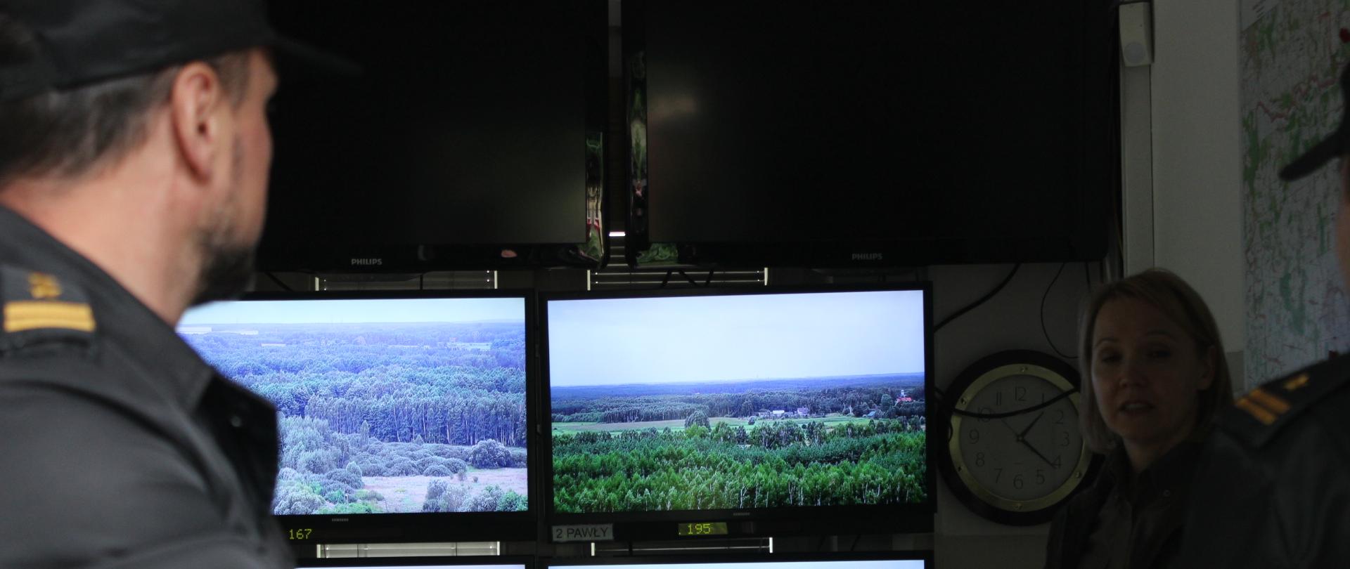 Przedstawiciele Państwowej Straży Pożarnej w punkcie alarmowo-dyspozycyjnym Nadleśnictwa Bielsk podczas prezentacji systemu wczesnego wykrywania dymu. W tle monitory, na których wyświetlany jest obraz z kamer monitoringu oraz przedstawiciel nadleśnictwa prowadzący prezentację.