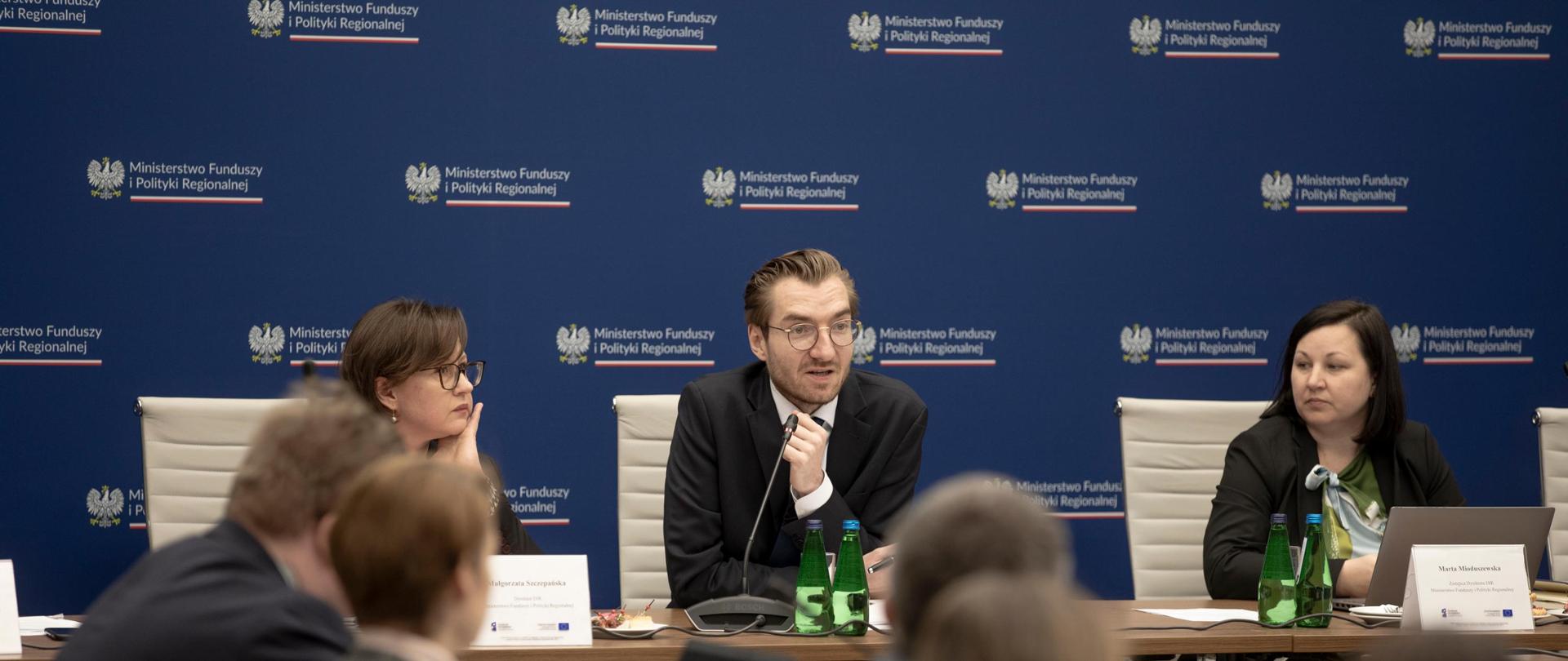 Trzy osoby siedzą przy stole. W środku, między dwiema kobietami, siedzi wiceminister Jan Szyszko. 