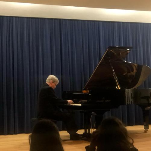 Na zdjęciu widać mężczyznę siedzącego przy fortepianie, z zamkniętymi oczami, co sugeruje głębokie zaangażowanie w grę. W tle niebieskie zasłony, co nadaje scenie profesjonalny wygląd. W dolnej części kadru widać sylwetki słuchaczy koncertu.