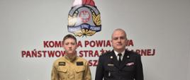 Komendant powiatowy Państwowej Straży Pożarnej stoi obok nowej funkcjonariuszki. Oboje pozują do zdjęcia na tle ściany z emblematem PSP.