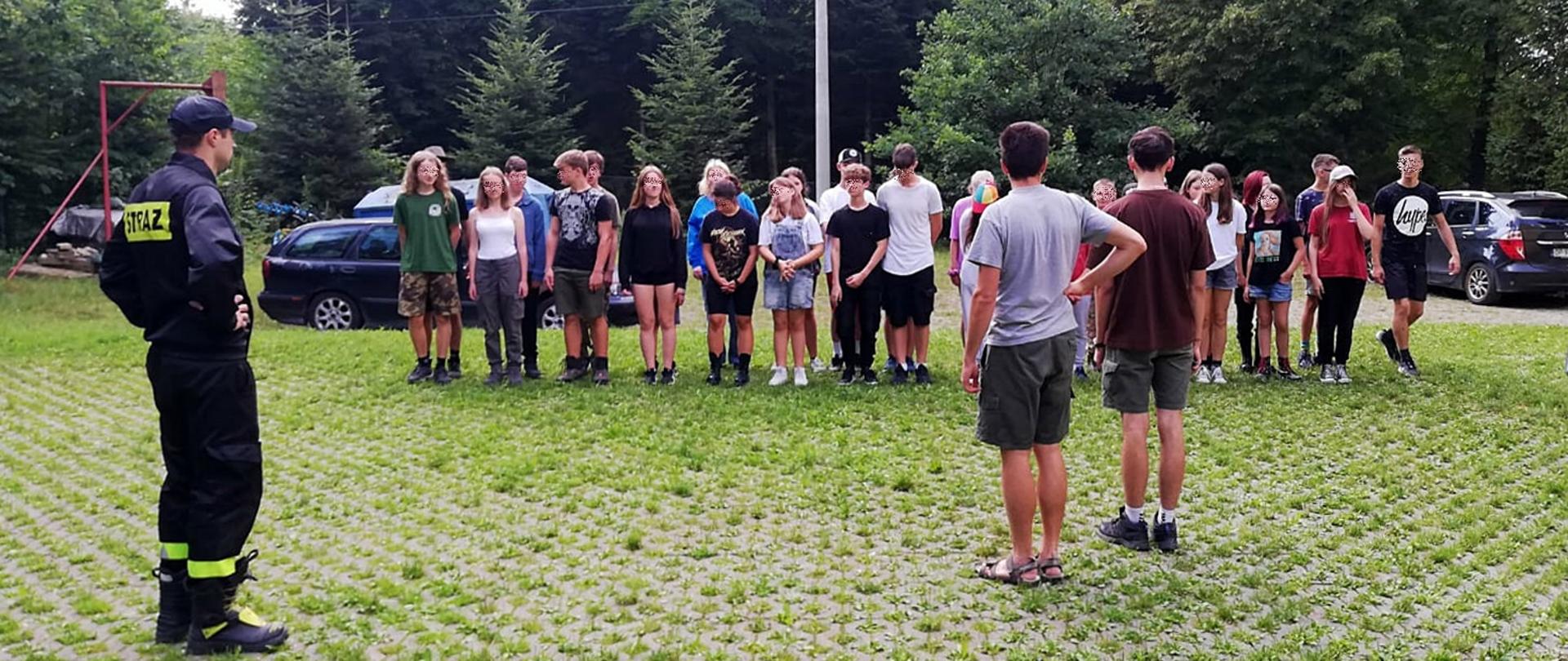 Widok z przodu. Strażak PSP widoczny po lewej stronie stoi przed harcerzami, którzy stoją w dwuszeregu. Przed frontem harcerzy stoi dwóch instruktorów ZHP i sprawdza stan osobowy.