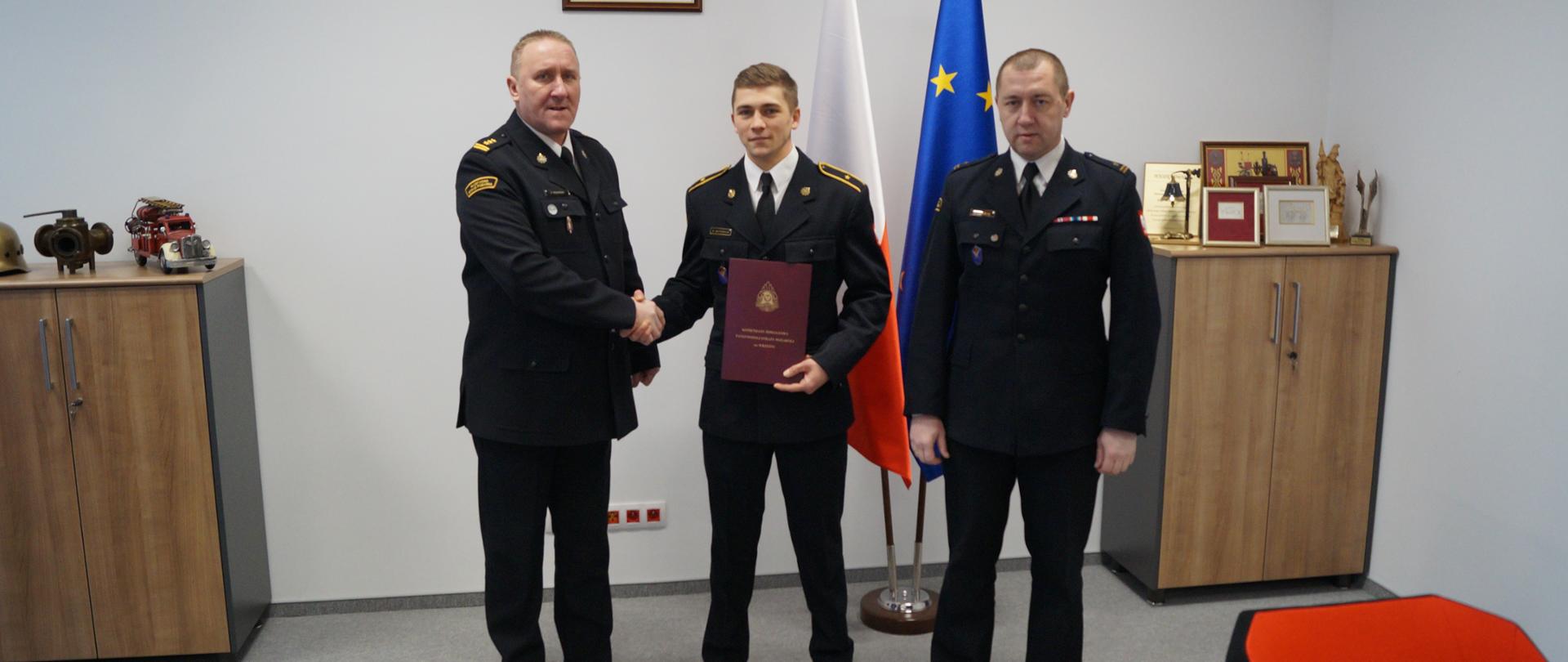Zdjęcie przedstawia trzech strażaków w mundurach wyjściowych stojących na tle godła państwowego, flagi Polski i Unii Europejskiej