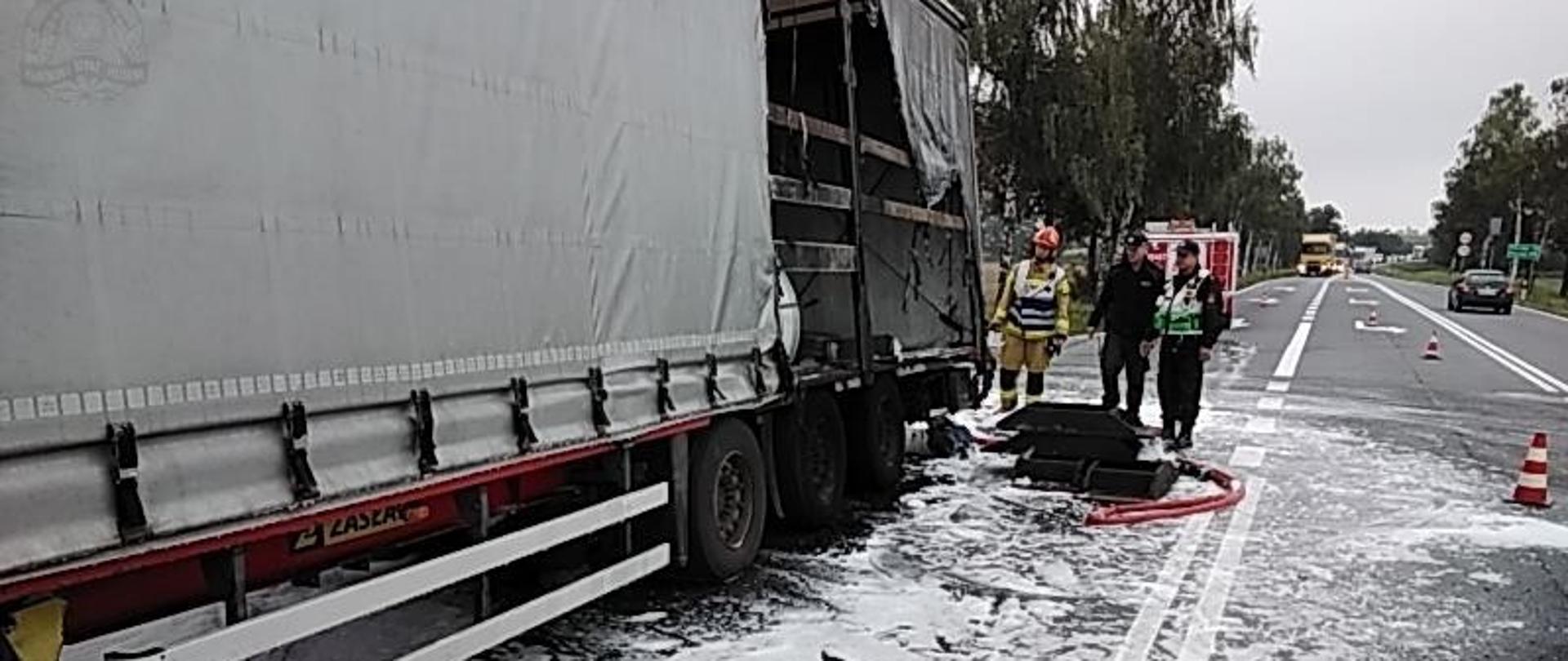 Droga asfaltowa pokryta pianą gaśniczą. Na niej stoi naczepa samochodowa, częściowa nadpalona od tyłu. Obok niej stoi dwóch strażaków i policjant. W tle widać samochód pożarniczy, zakorkowany pas drogi oraz drzewa.