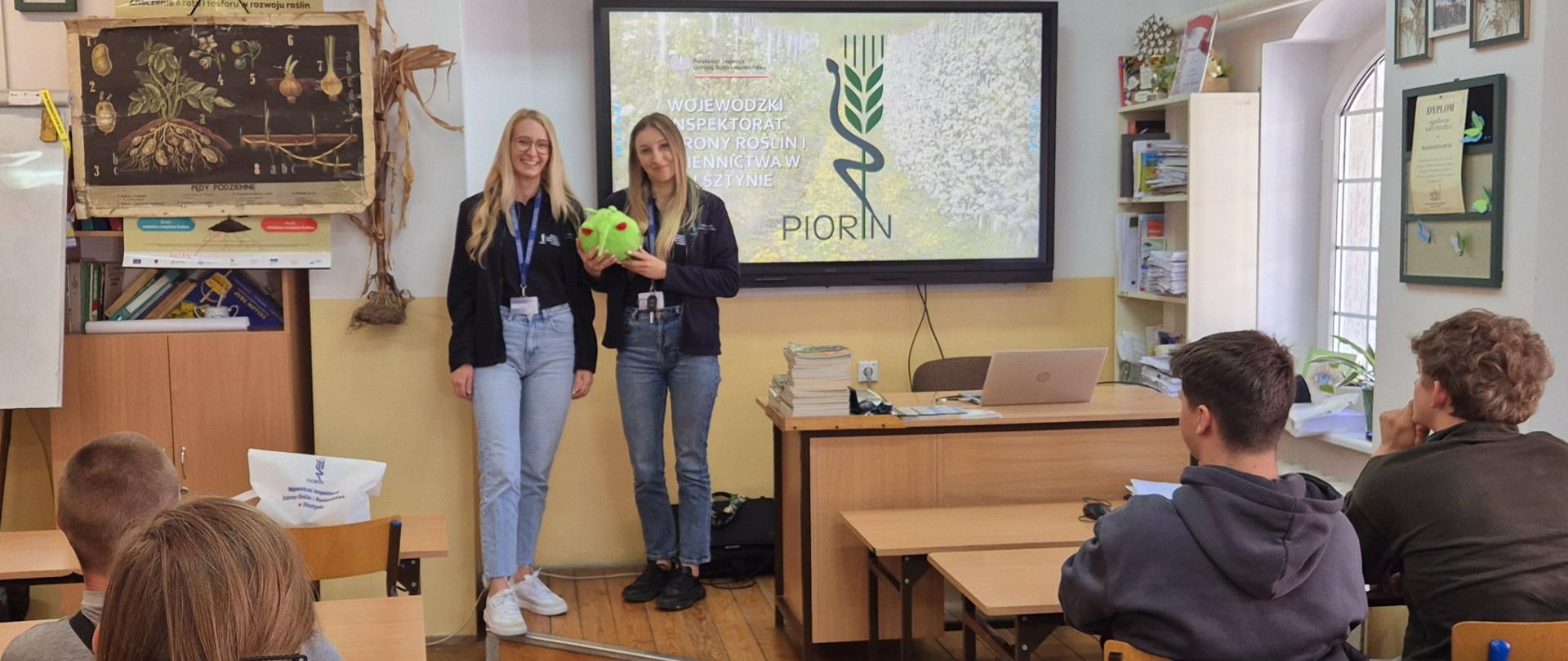 Dwie kobiety w granatowych bluzach stoją przed tablicą na której widnieje napis PIORIN. Jedna z
kobiet trzyma zieloną maskotkę z czerwonymi oczami. Przed nimi grupa słuchaczy siedząca w szkolnych ławkach