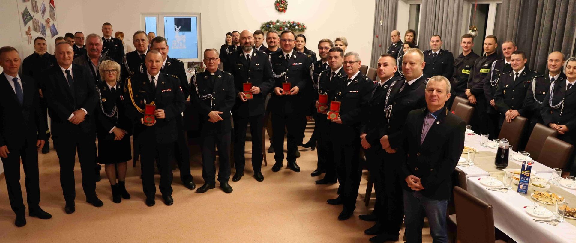 Wewnątrz pomieszczenia stoi duża grupa strażaków w mundurach galowych, pozują do zdjęcia, spotkanie świąteczne. Z prawej strony widać stół z białym obrusem.