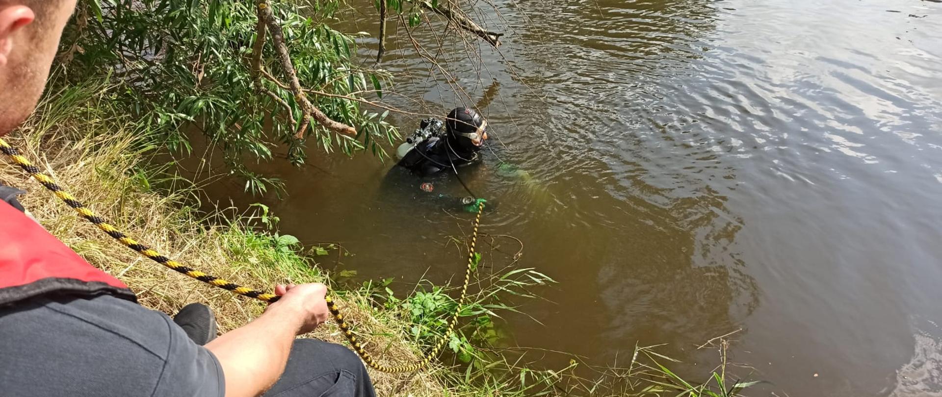 Na zdjęciu z lewej częściowo widoczny strażak siedzi na trawie, asekuruje linką nurka w wodzie.